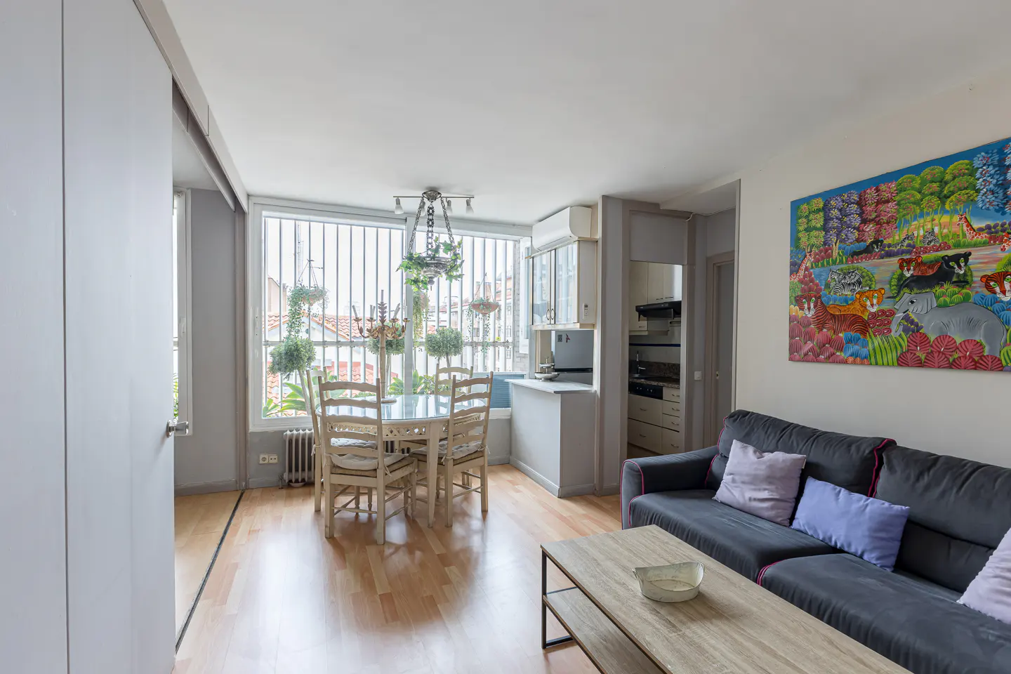 Bright apartment interior with wood floors, a gray sofa with pillows, and a round dining table near a window with hanging plants. A colorful painting hangs on the wall.