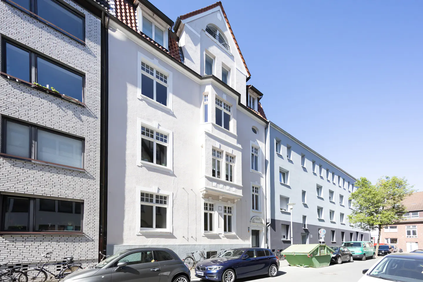 Street view of a multi-story apartment building with white and gray facades, cars parked along the curb, and a clear blue sky.