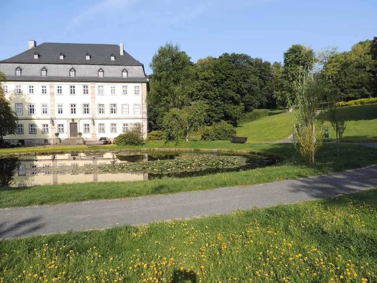 A large, cream-colored building with a dark roof is reflected in a pond with lily pads, surrounded by green grass and trees.
