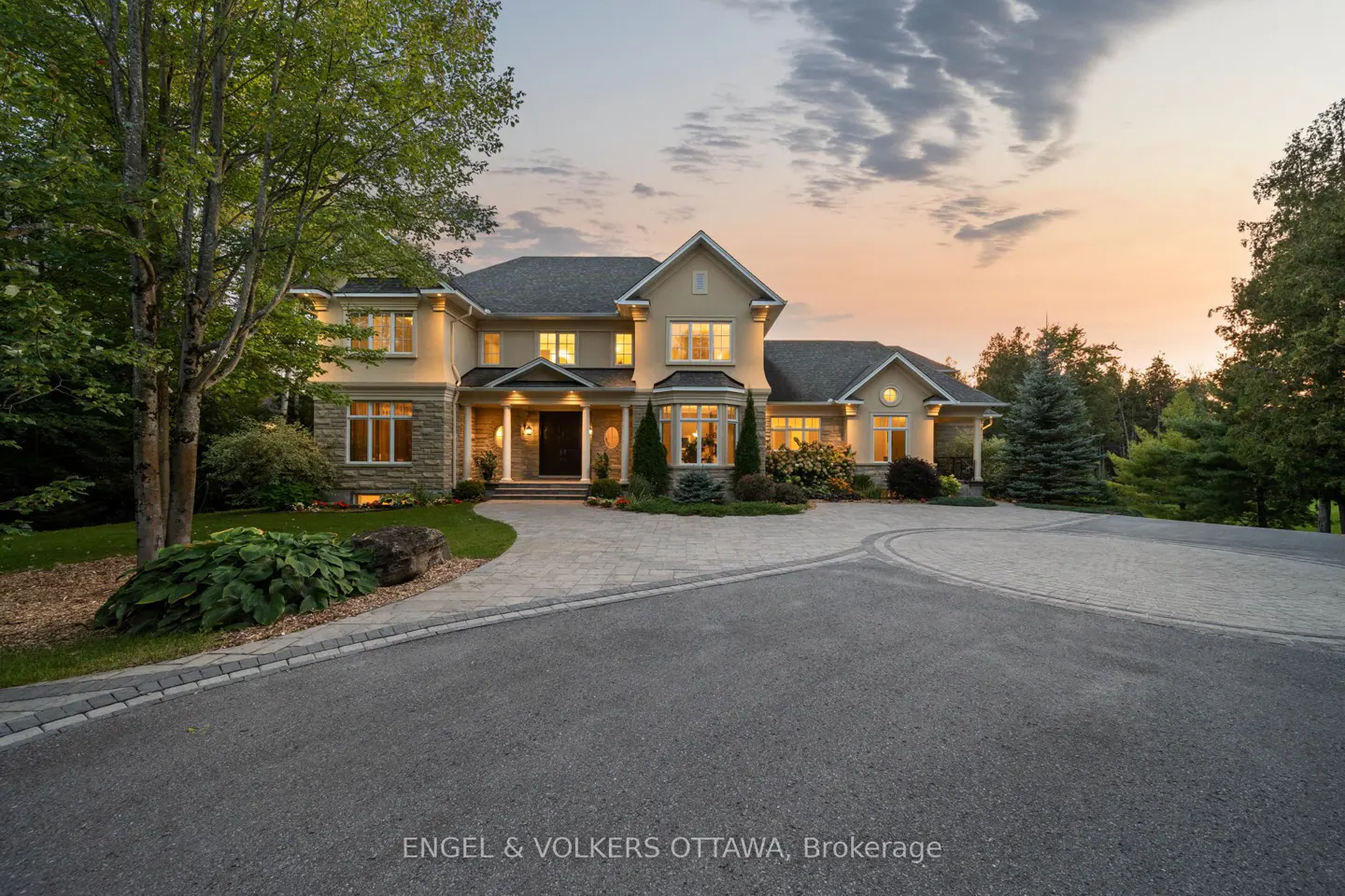 Exterior view of a large, two-story beige house with a gray roof and stone accents, set against a sunset sky.