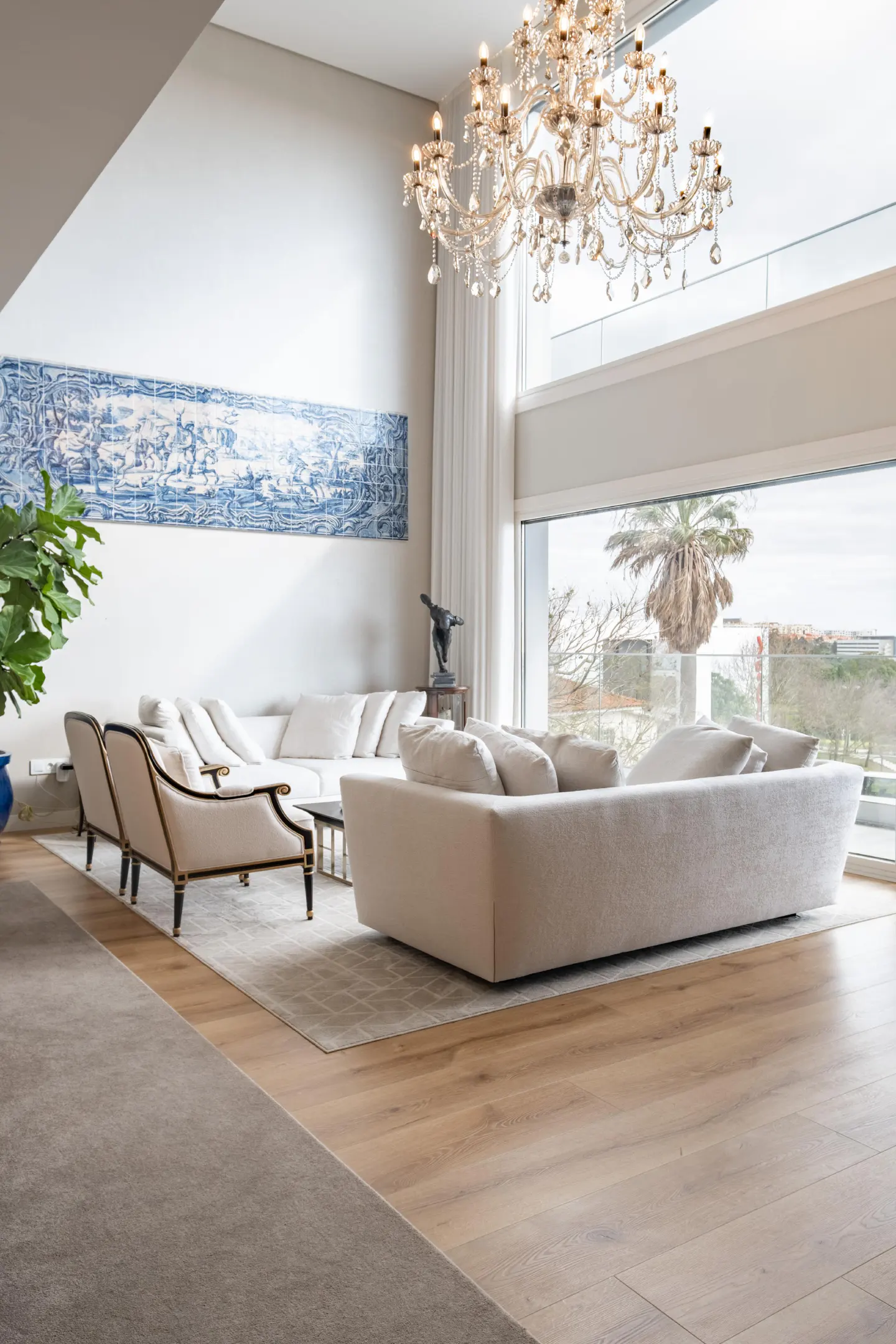 Bright living room with a large window, a crystal chandelier, and white sofas. A blue tile mural adorns the wall.