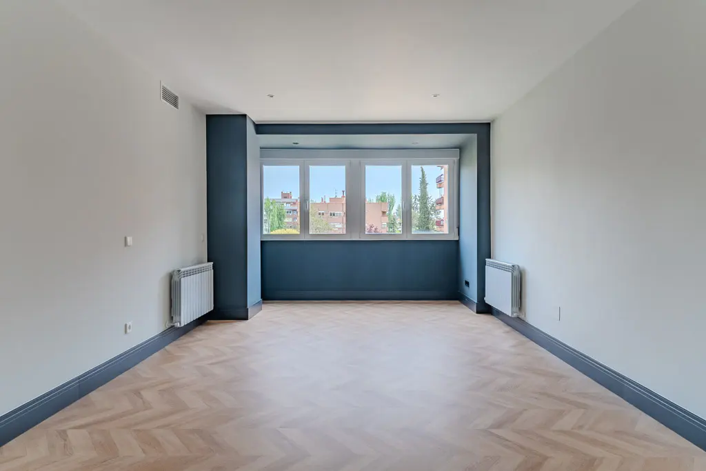 Empty room with herringbone wood floors, white walls, and a blue accent wall with four windows. Radiators are mounted on the walls.