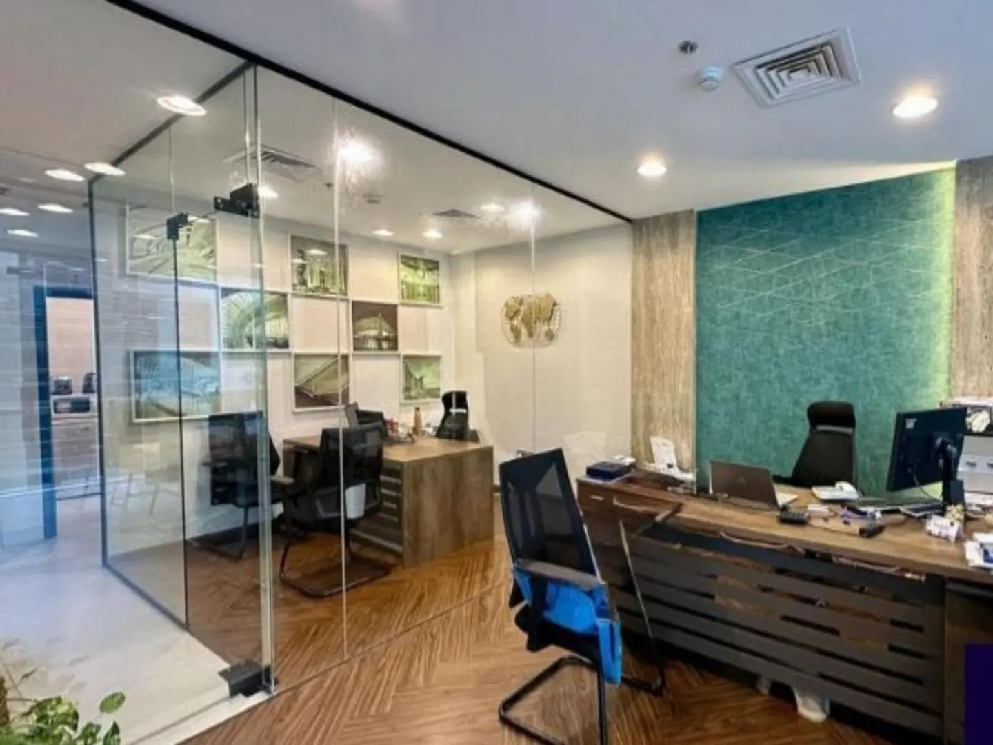 Office space with glass walls, wooden floors, and desks. Artwork adorns the walls, and chairs are positioned at the desks.