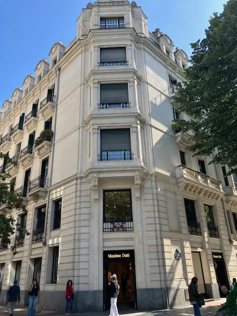 Exterior of a light-stone building with a Massimo Dutti store at street level. Balconies and trees are visible.