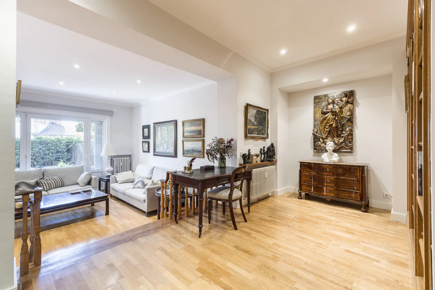 Bright living room with light wood floors, white walls, and recessed lighting. Sofas, a dark wood table, and art adorn the space.