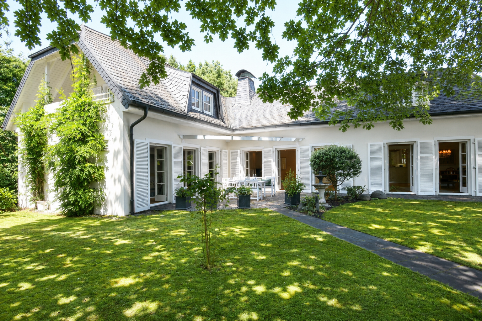 White house with a slate roof, white shutters, and a patio set in a lush green backyard with a stone path.
