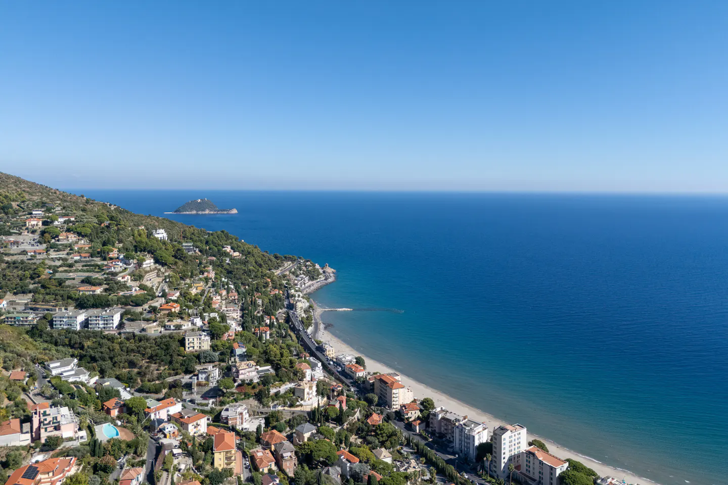 Aerial view of a coastal town with houses on a hillside, a sandy beach, and blue ocean under a clear sky.