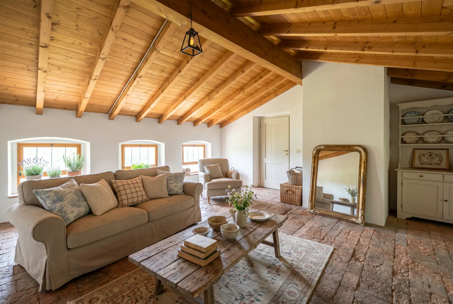 Attic living room with exposed wood beam ceiling, brick floor, beige sofa, and rustic wood coffee table. Natural light from windows.