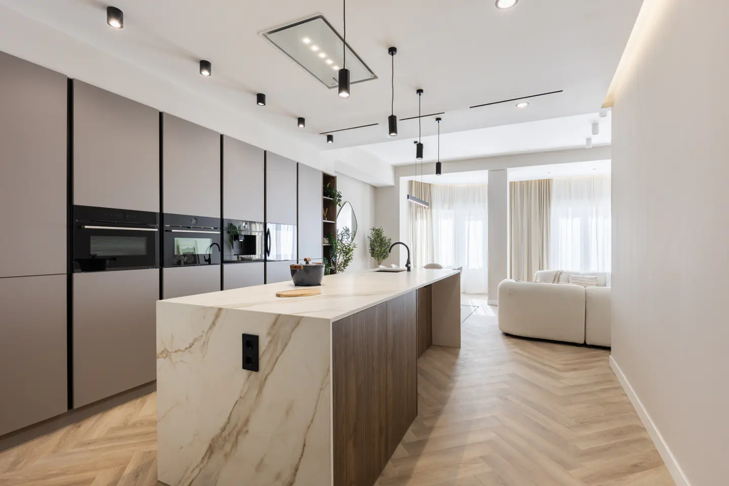 A modern kitchen with gray cabinets, a marble island, and herringbone wood floors. Black pendant lights hang above the island. A white sofa is visible in the background.