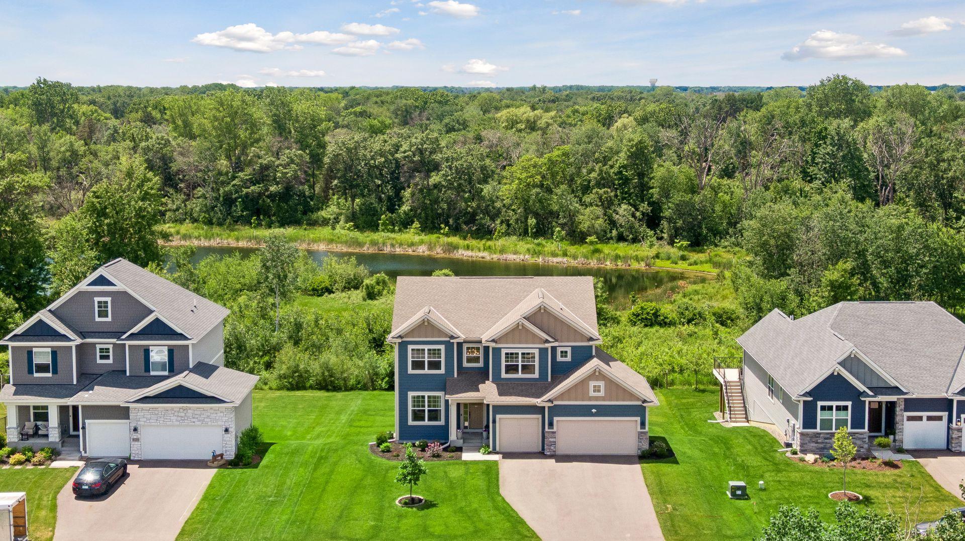 Aerial view of three modern suburban homes with blue siding and two-car garages, situated before a pond and dense forest.