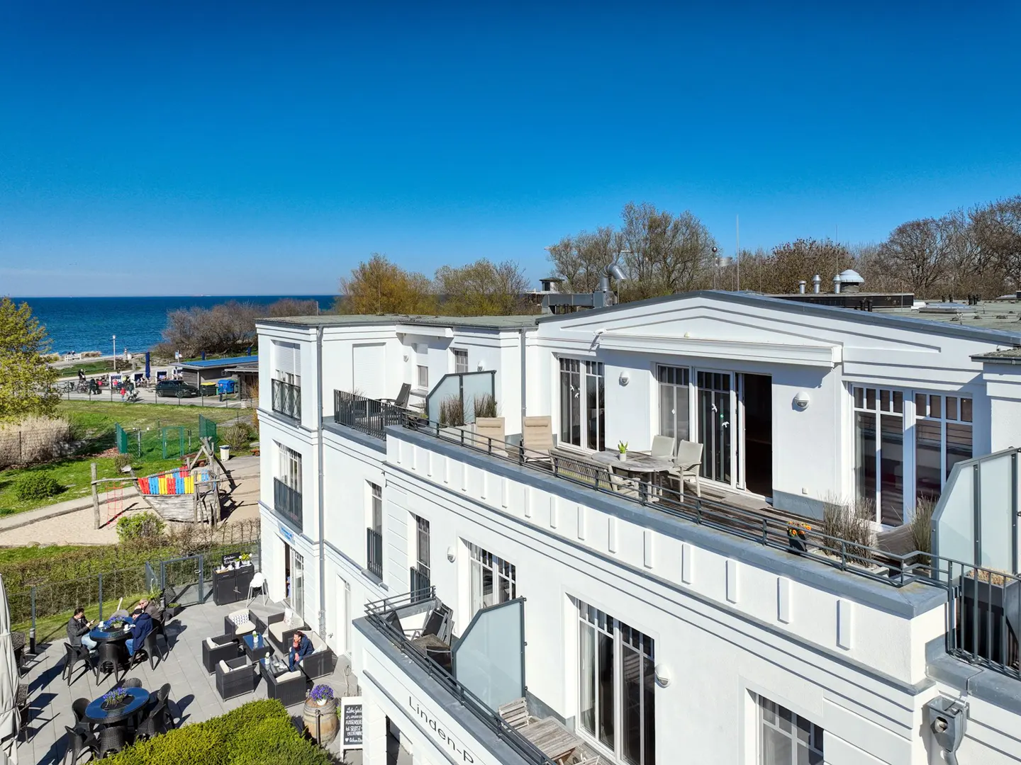 View of a white building with balconies overlooking the sea and a park with a playground. People are seated at tables on a patio.