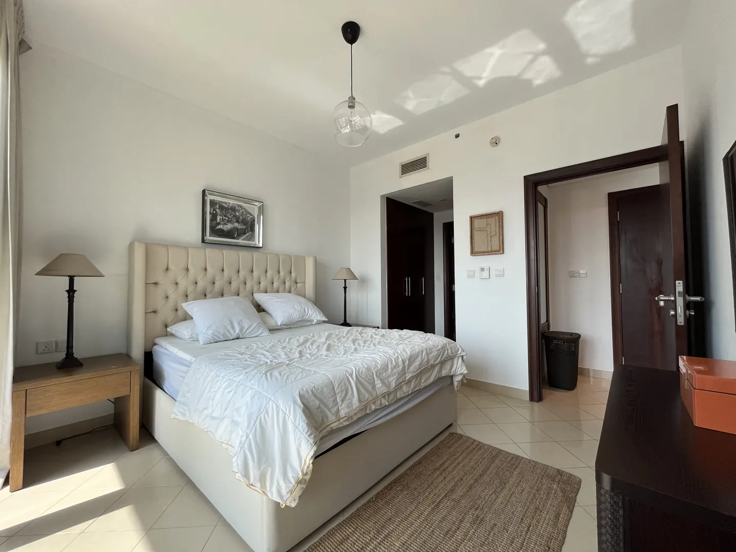 Bedroom with a tufted headboard bed, white bedding, side tables with lamps, and a brown rug on a tiled floor.
