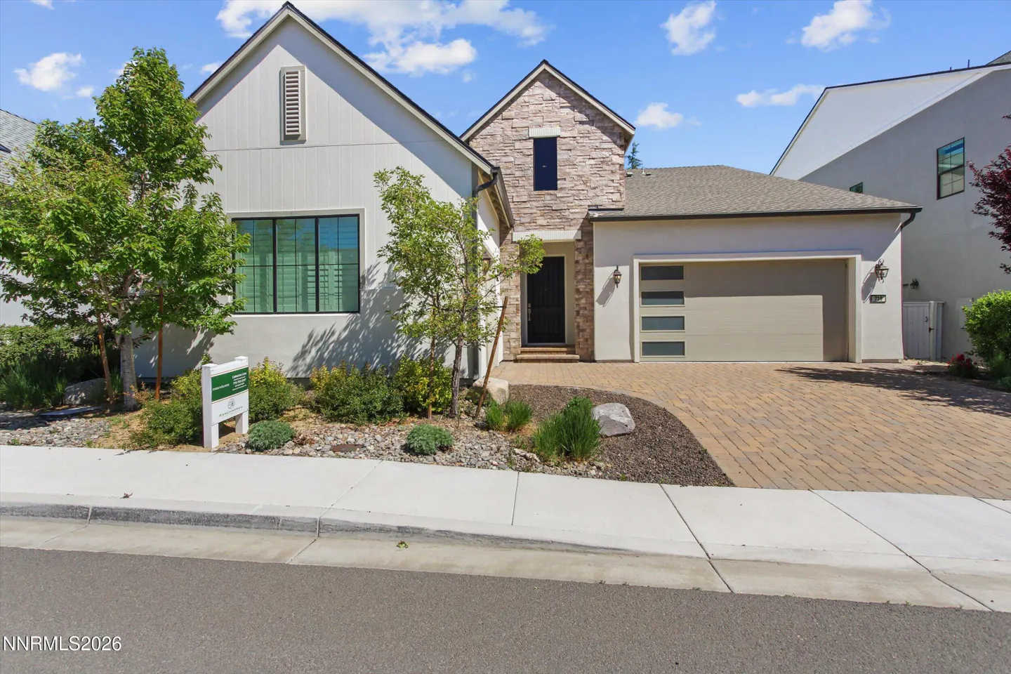 A modern two-story house with a stone facade, a gray garage door, and a brick driveway on a sunny day.