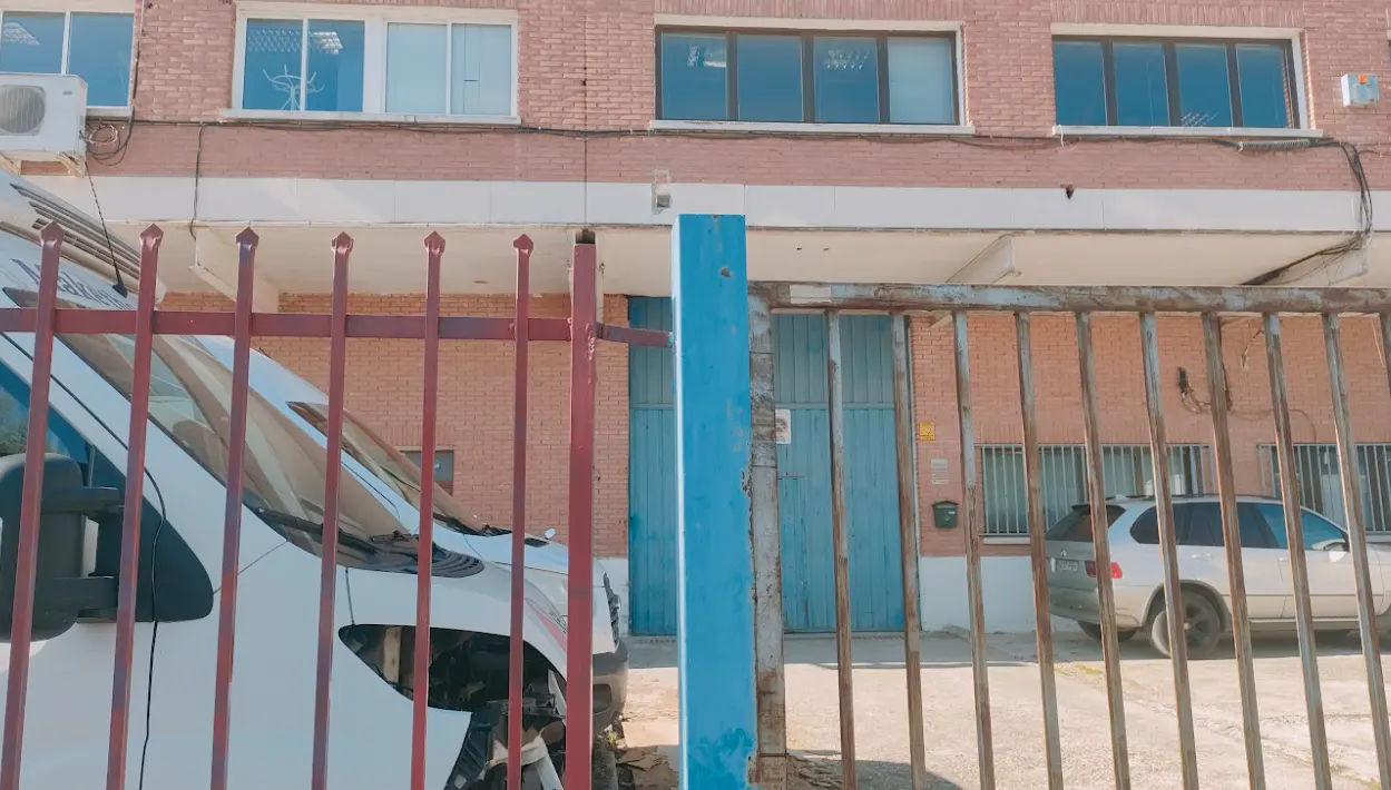 Exterior of a brick building with a metal gate, a white van, and a silver SUV visible through the gate.
