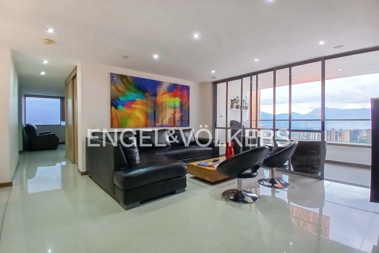 Bright living room with black leather sectional, chairs, and colorful art. Large windows offer a city view. Engel & Völkers logo visible.