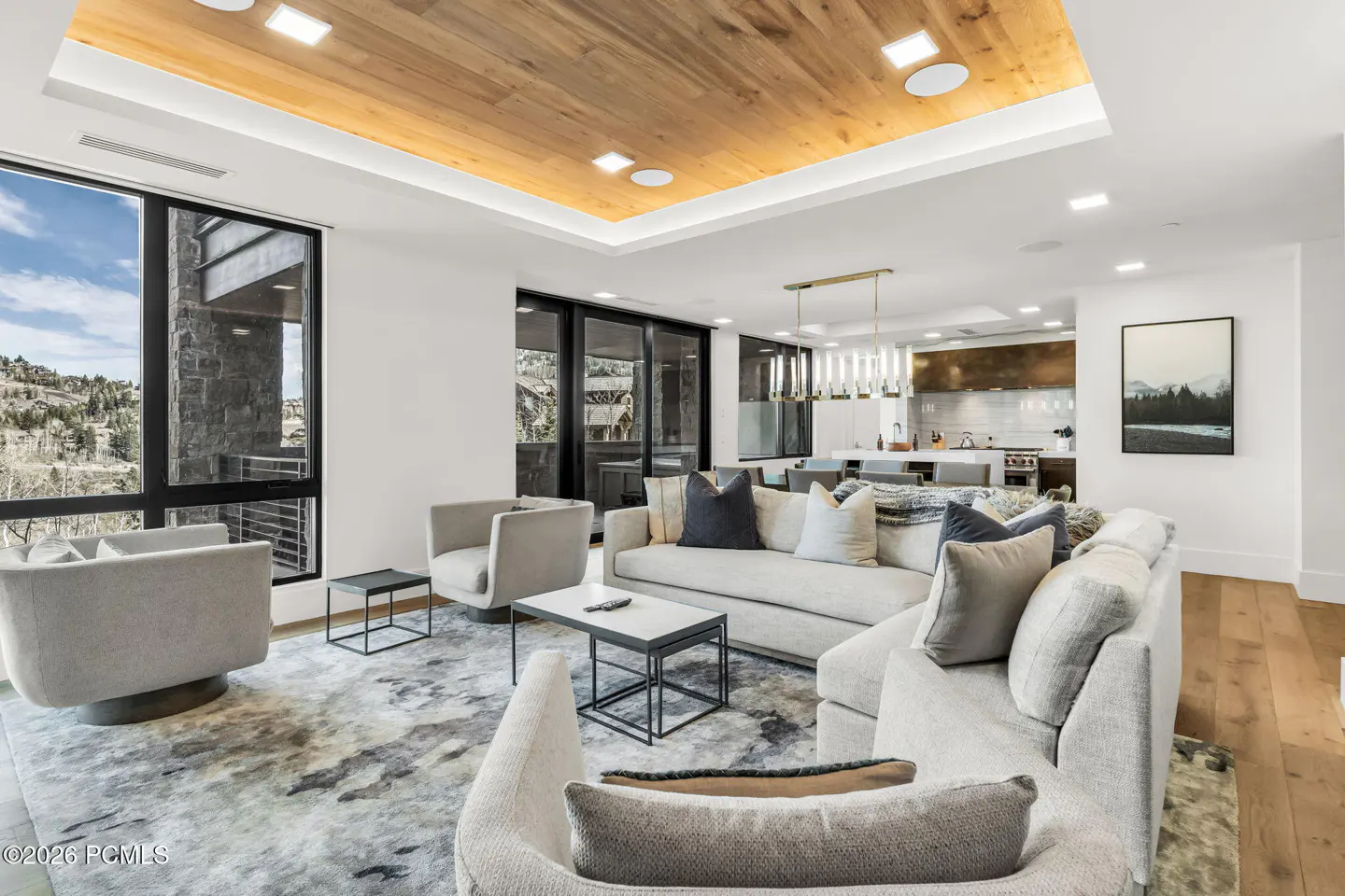 Bright, modern living room with gray sofas, chairs, and a patterned rug. Large windows offer a mountain view. A kitchen is visible in the background.