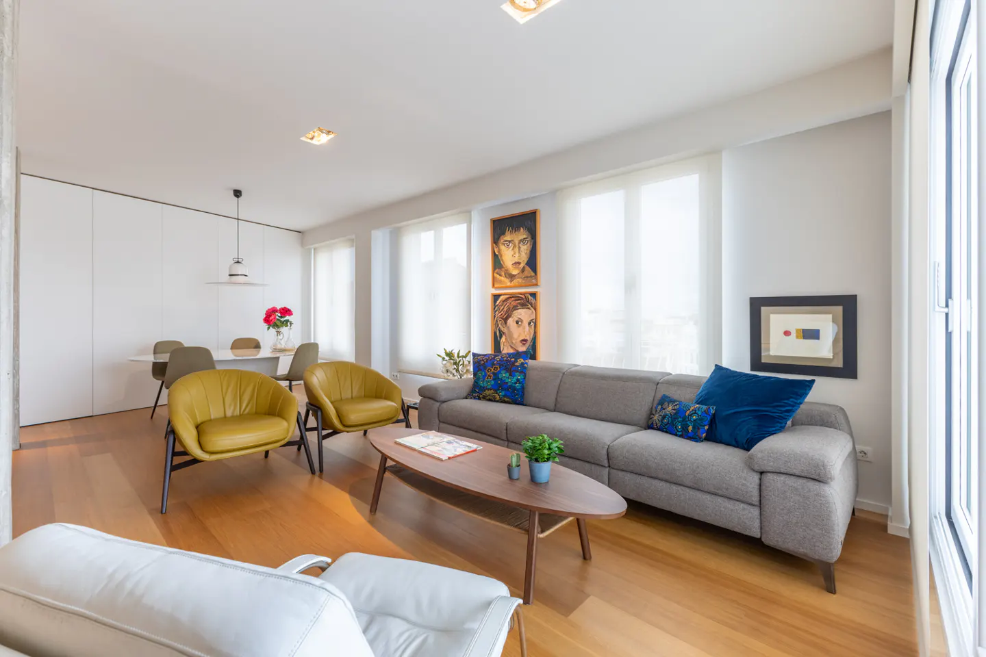 Bright living room with wood floors, grey sofa with blue pillows, two yellow chairs, and a wood coffee table. Art on the walls.
