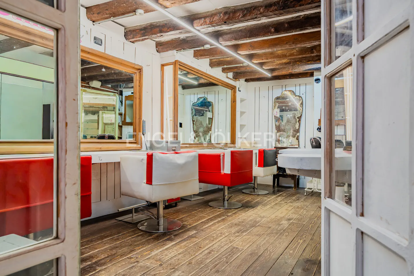 Interior view of a salon with red and white chairs, wooden floors, and large mirrors. The ceiling has exposed wooden beams and a modern light fixture.