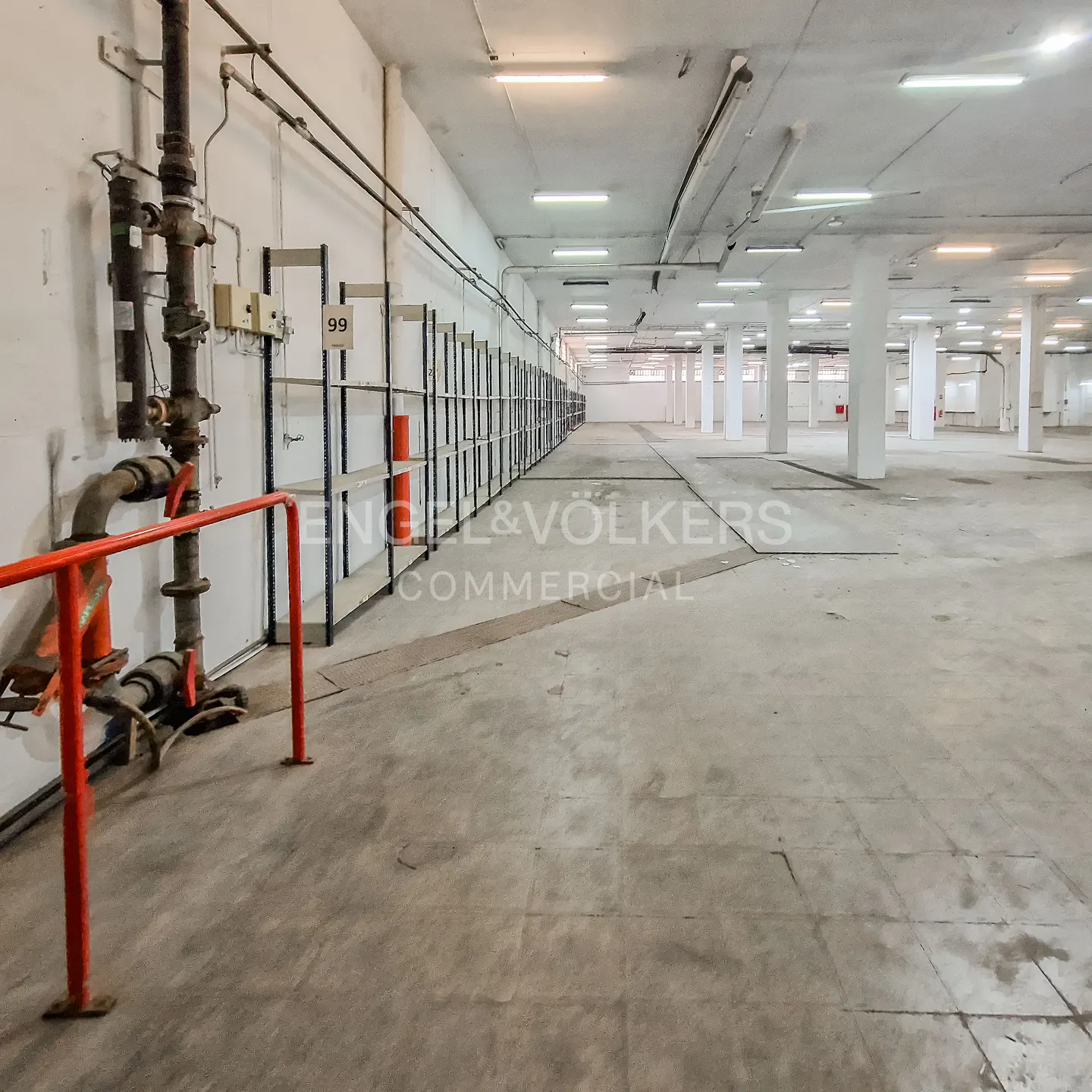 Wide shot of a large, empty warehouse with concrete floors, white walls, and rows of metal shelving. Overhead lighting illuminates the space.
