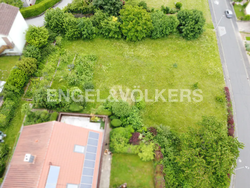 Aerial view of a grassy lot for sale, bordered by trees and a road. A house with solar panels is visible in the lower left. "Engel & Völkers" is overlaid.