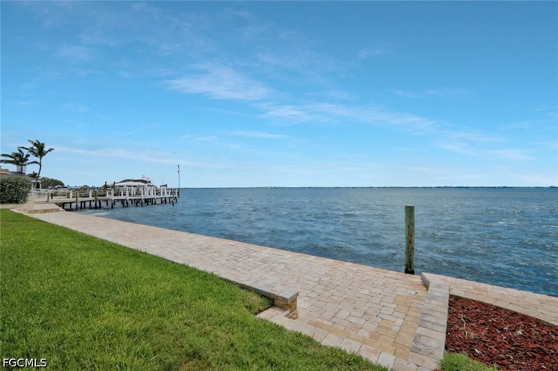 Waterfront property view with a stone walkway, green grass, and a dock extending into the blue water under a bright sky.