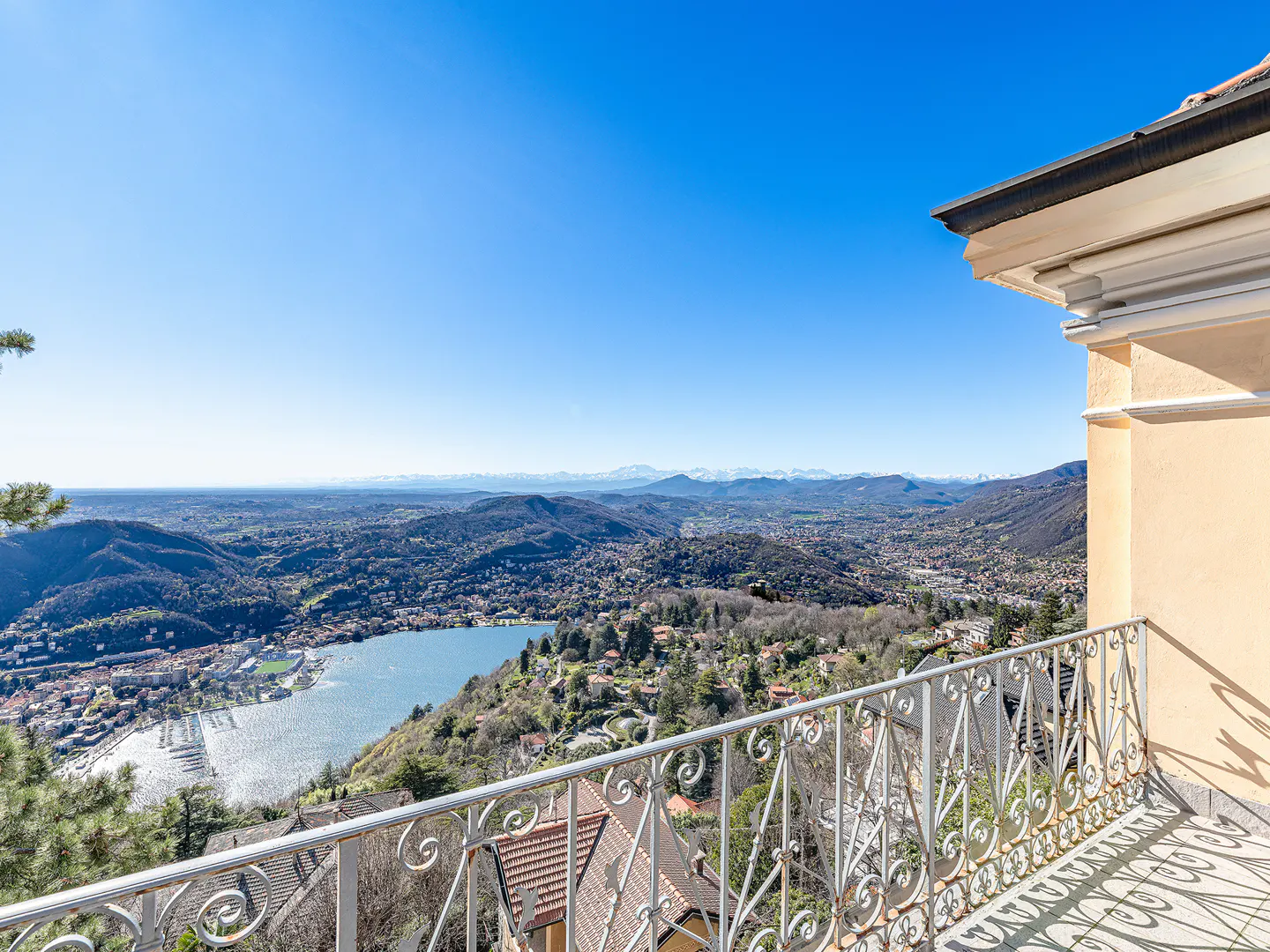 View from a balcony with white railings overlooking a lake, mountains, and a town under a clear blue sky.