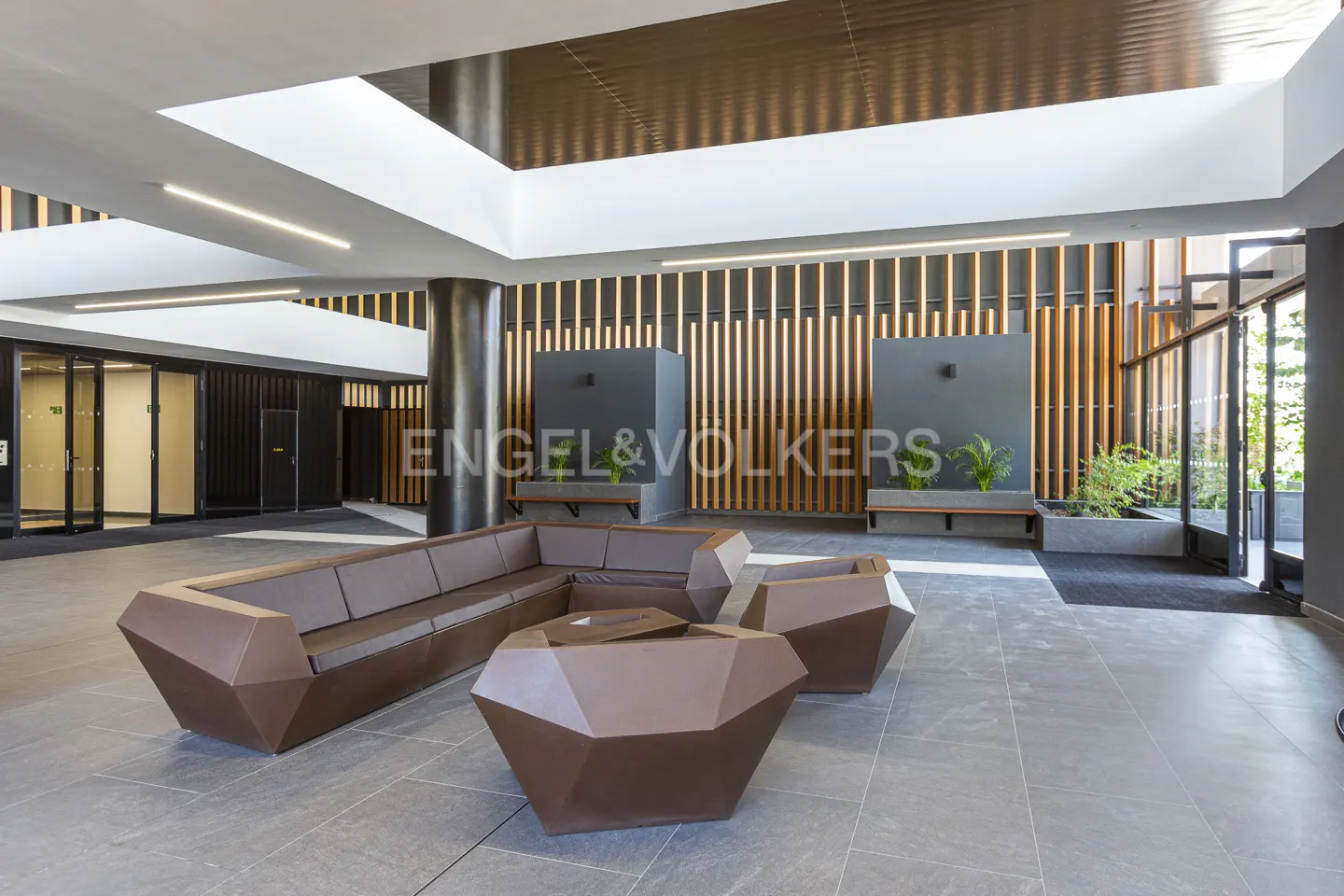 Lobby area with brown geometric sofa and table. Engel & Volkers logo on the wall with plants. Gray tile floor and modern lighting.