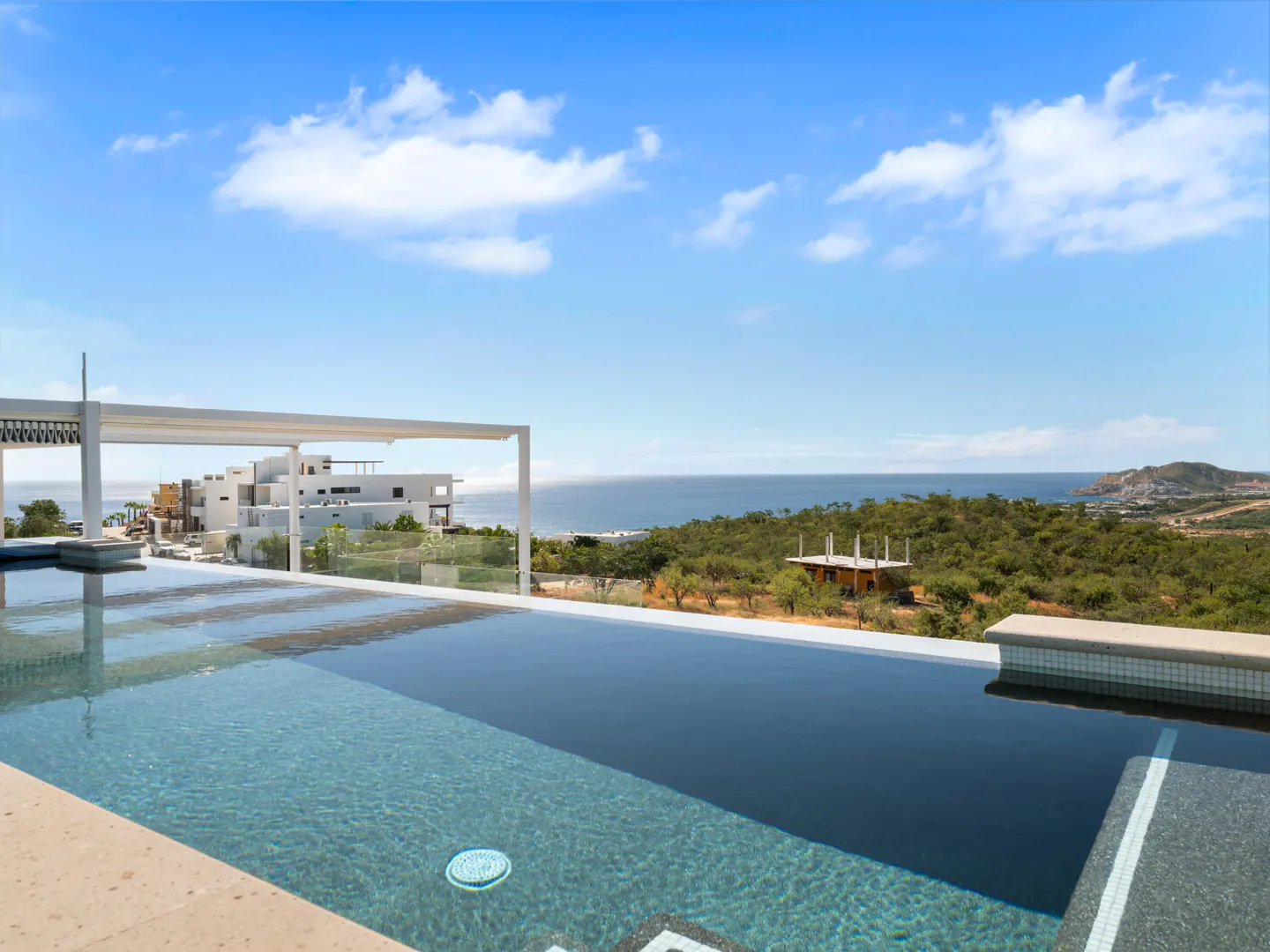Infinity pool with blue water overlooking the ocean and green hills under a blue sky with white clouds.