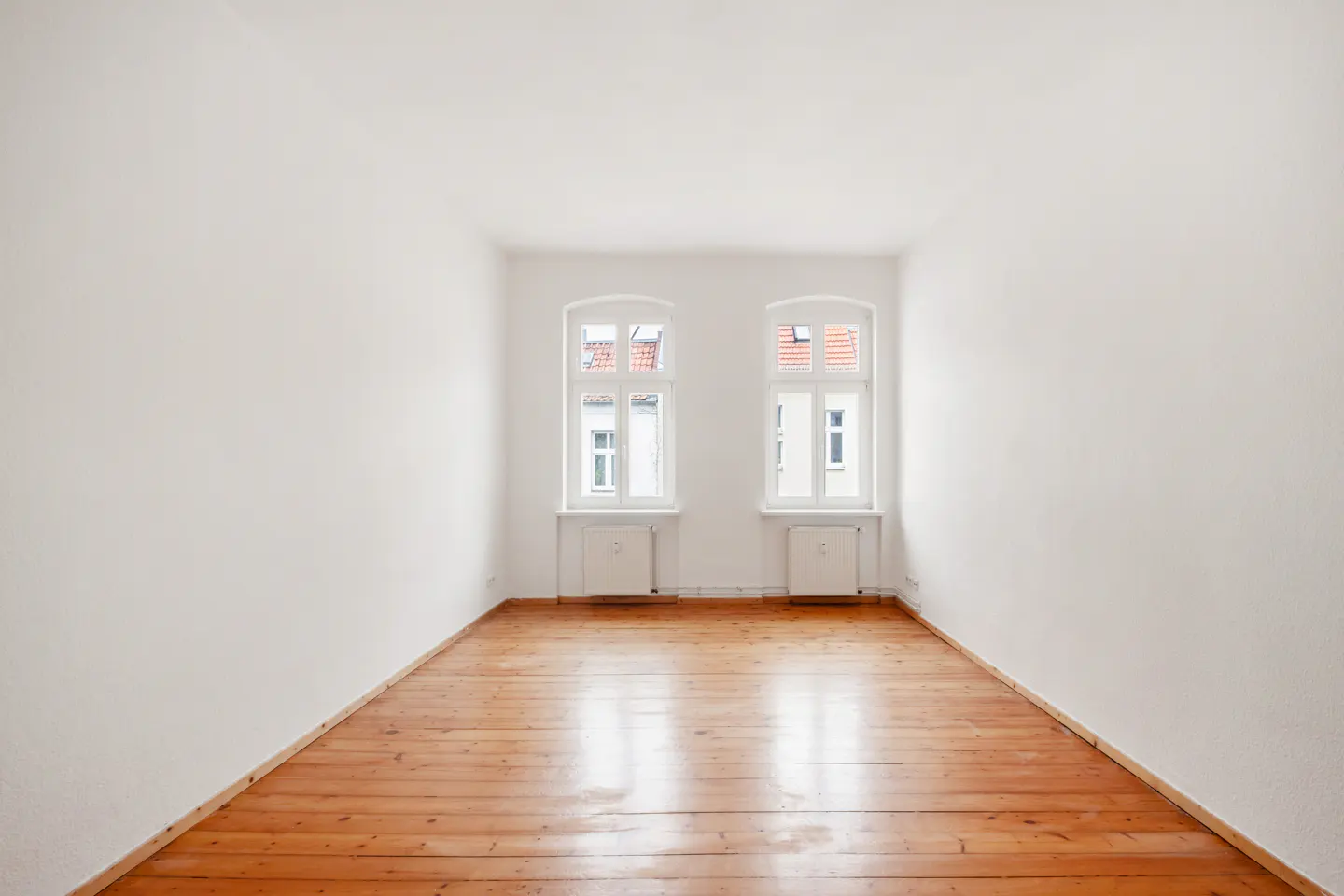 Empty room with white walls, wood floor, and two windows. Radiators are under each window.
