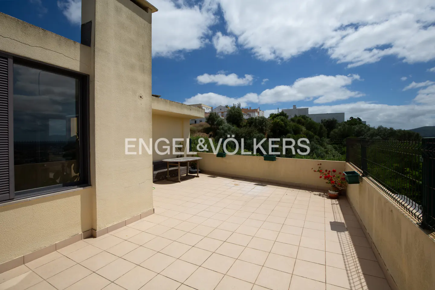 A sunny terrace with beige tiles, a wooden table, and a view of green hills under a blue sky. The Engel & Völkers logo is superimposed.