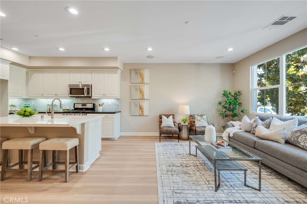 Open-concept kitchen with white cabinets and island, adjacent to a living area with a grey sofa and patterned area rug. [AI-generated text]