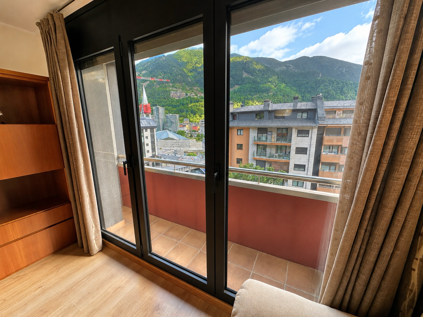 View from a room through large glass doors onto a balcony overlooking a town with mountains in the background.
