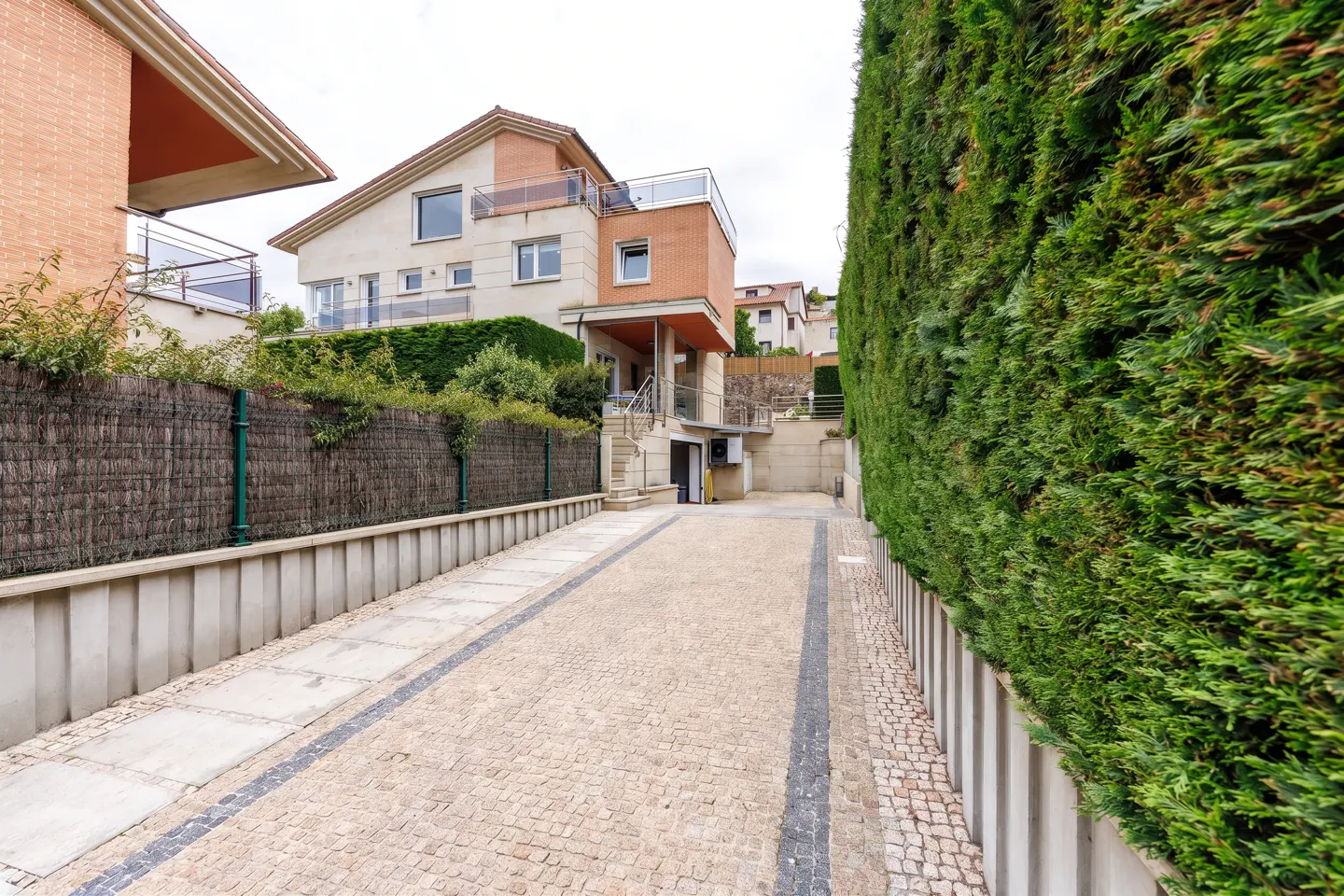Exterior view of a beige stone driveway leading to a modern house with a green hedge on the right.