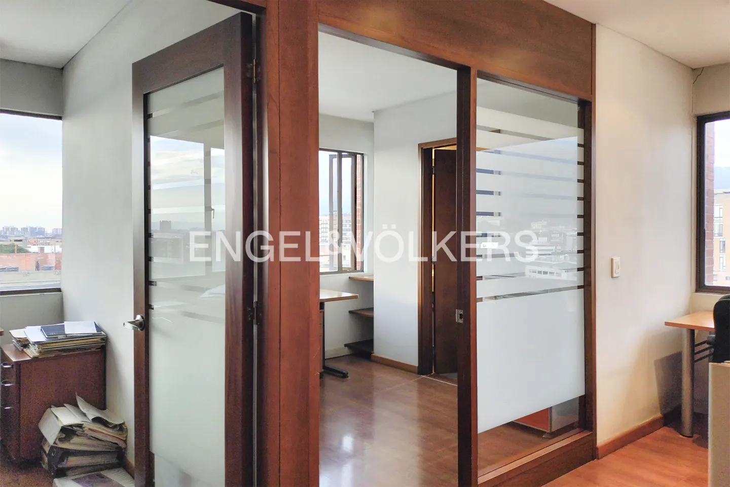 Office interior with wood-framed glass doors, light walls, and wood floors. Desks are visible near windows with city views.