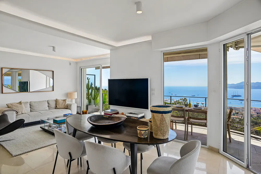 Bright living room with a dark wood table, white chairs, and a view of the ocean from the balcony.