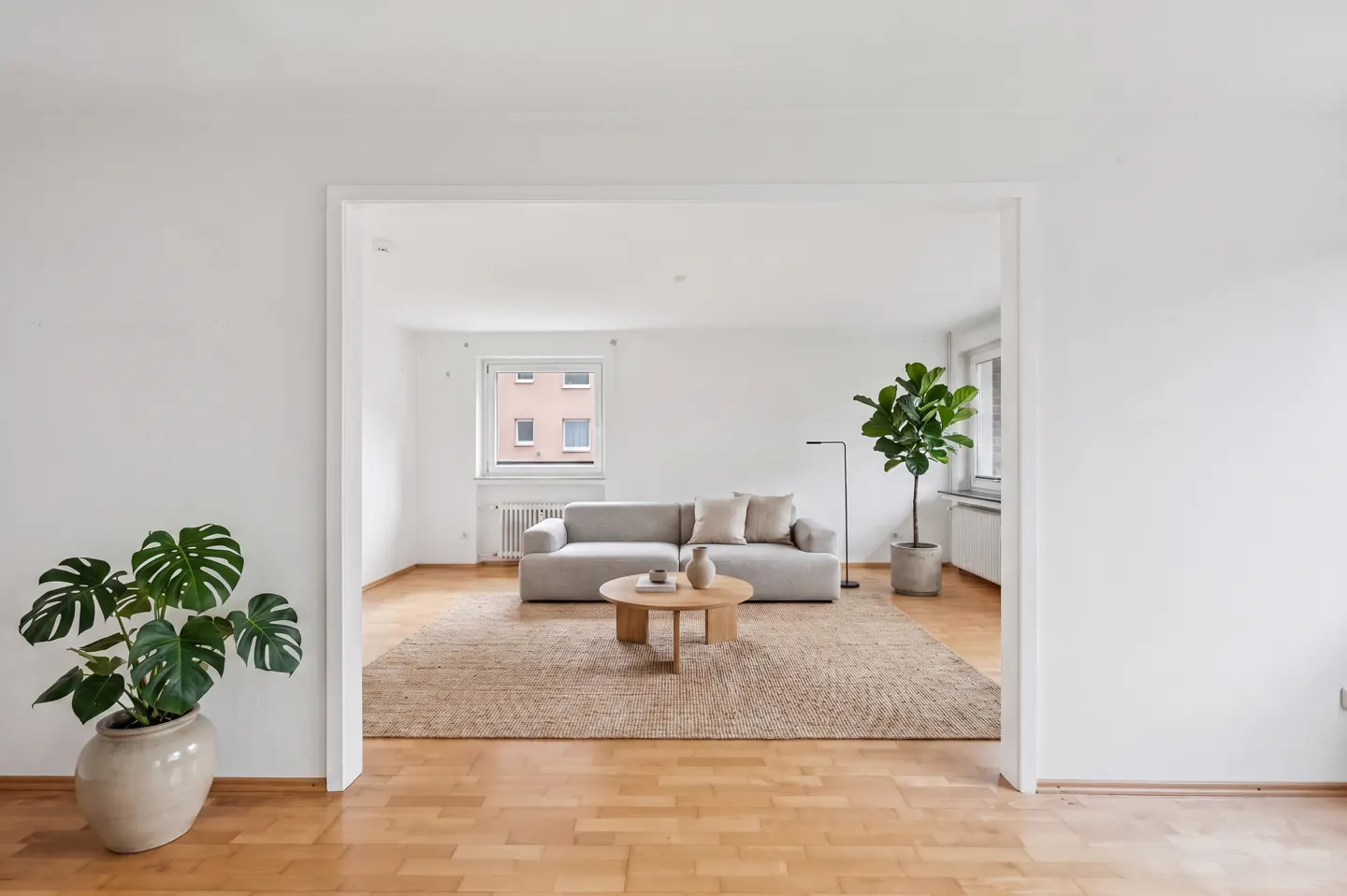 Bright, airy living room with hardwood floors. A gray sofa sits on a jute rug, with a round coffee table. A potted monstera plant is in the foreground.