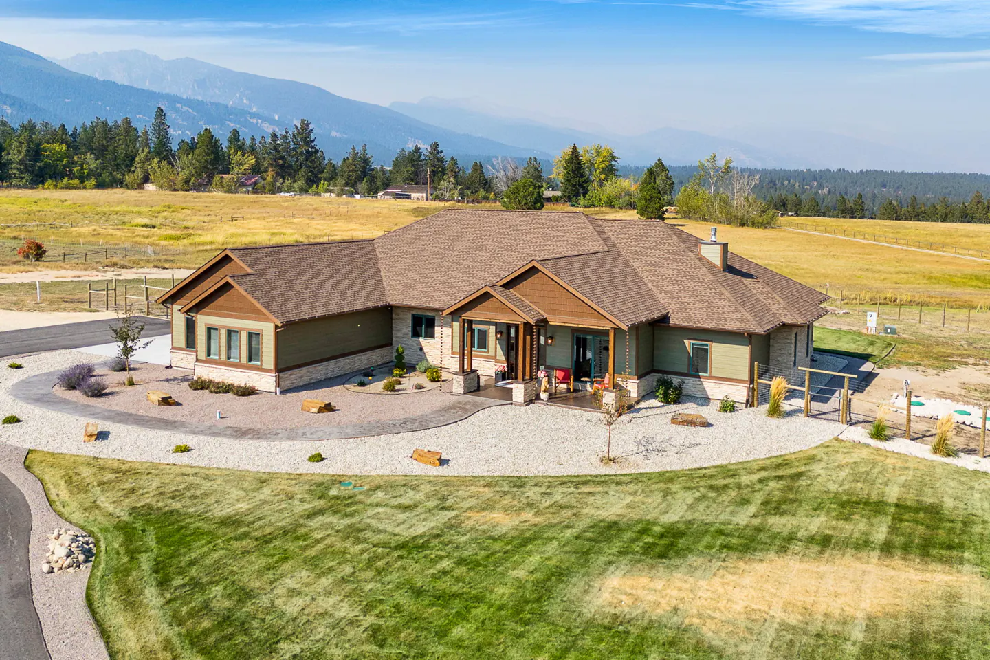 Aerial view of a green and stone house with a brown roof, surrounded by a green lawn and mountains in the background.
