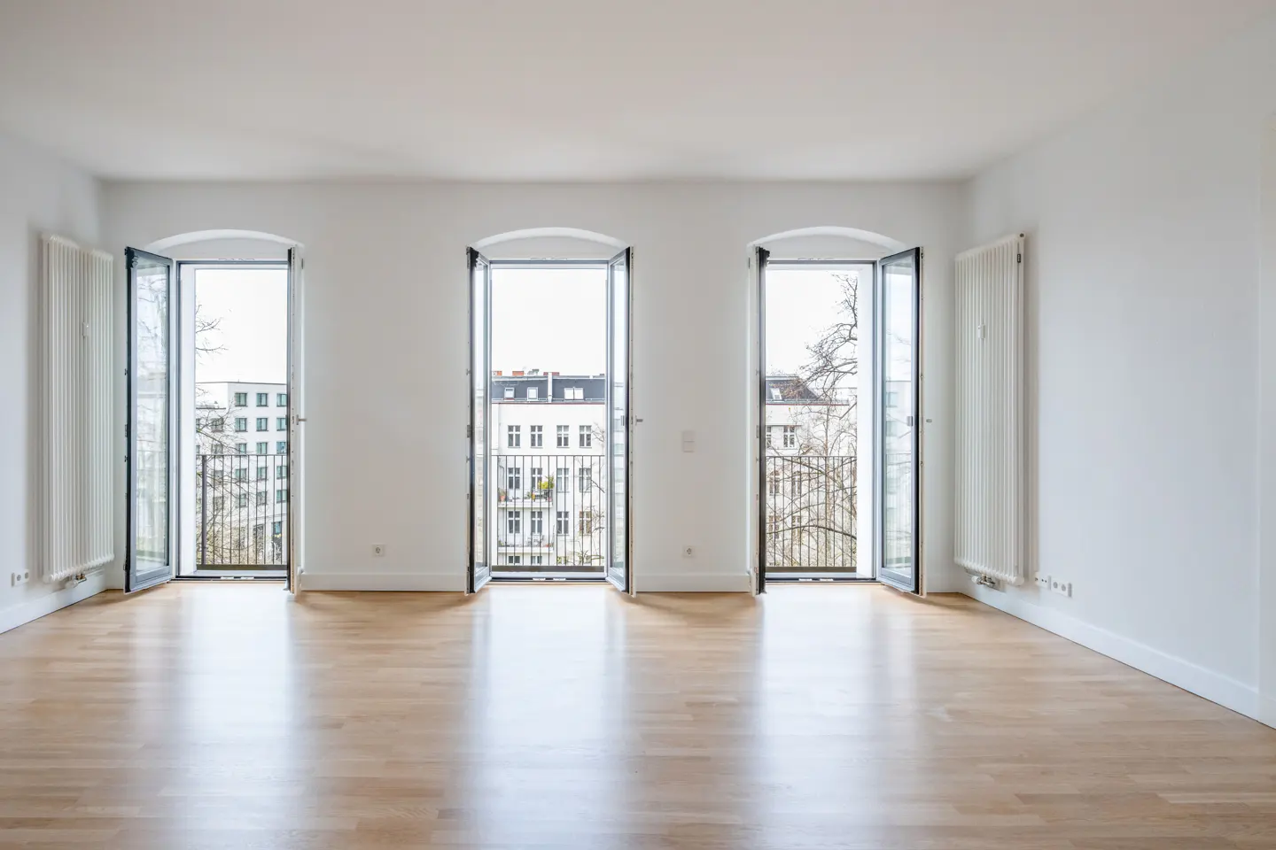 Bright, empty room with light wood floors and white walls. Three open, arched windows reveal city views. Radiators are mounted on the walls.