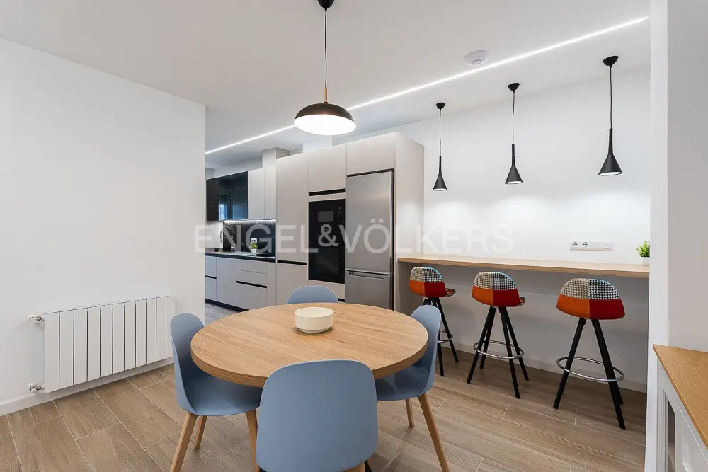A modern kitchen and dining area with a round wooden table, blue chairs, and colorful bar stools.
