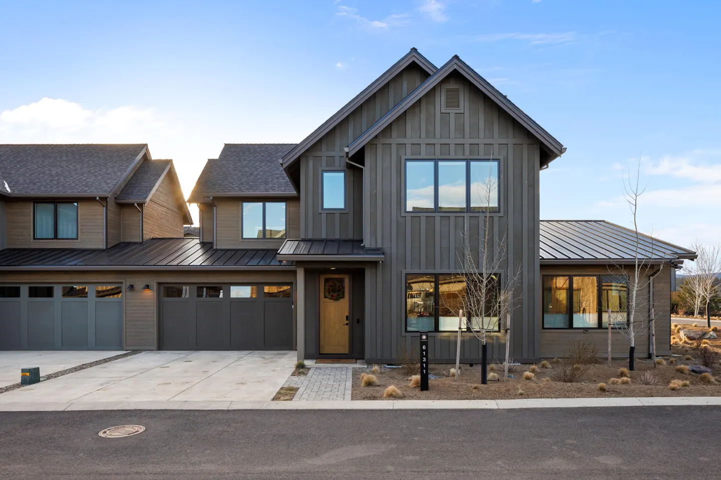 Two-story modern farmhouse with gray siding, black roof, and a light wood front door. A driveway leads to a three-car garage.