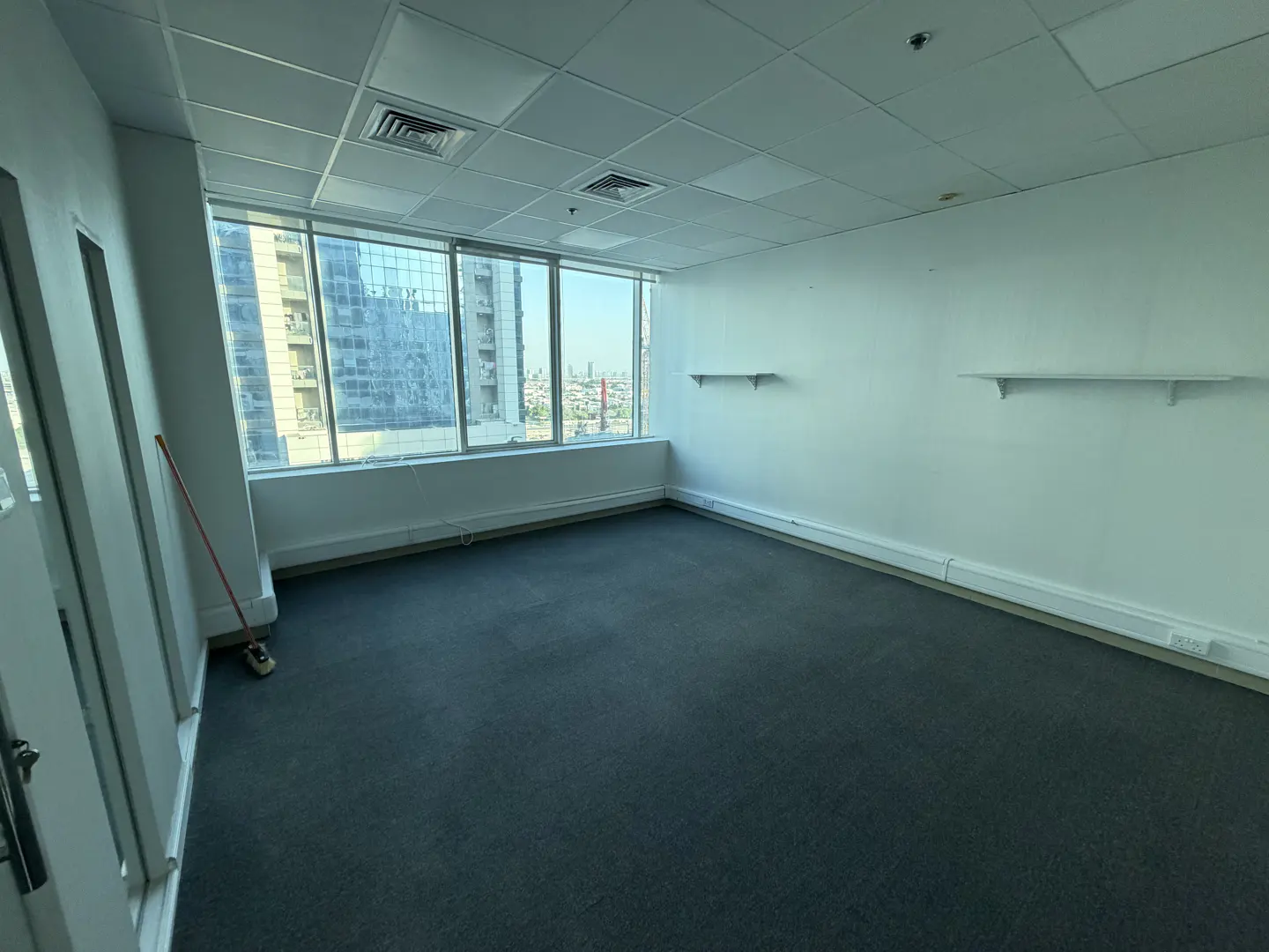 Empty office space with gray carpet, white walls, and large windows showing a city view. Two shelves are mounted on the wall.