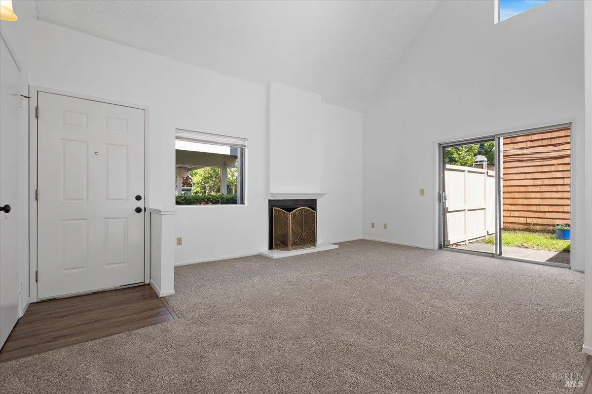 Bright, empty living room with white walls, beige carpet, and a fireplace. A sliding glass door leads to a patio.