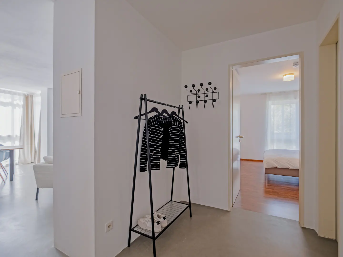Hallway with a black coat rack holding a striped jacket and shoes. A bedroom is visible through an open doorway.
