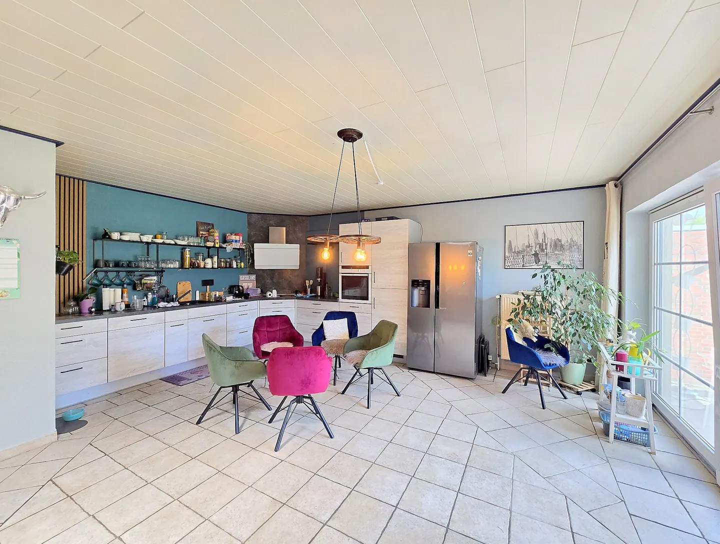 Modern kitchen with white cabinets, teal accent wall, open shelving, stainless fridge, and colorful velvet dining chairs.
