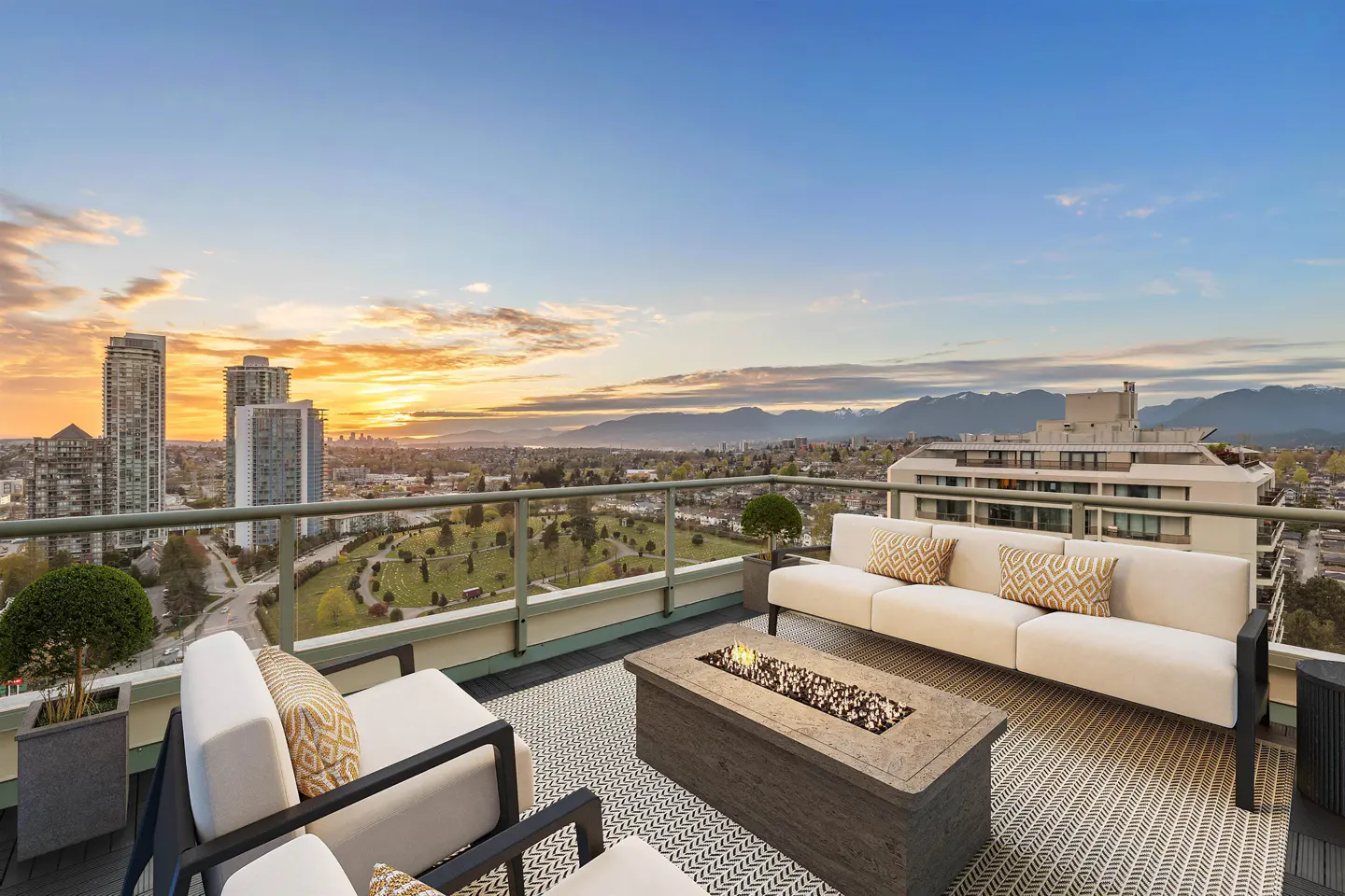A rooftop patio with white furniture, a fire pit, and a city view at sunset.