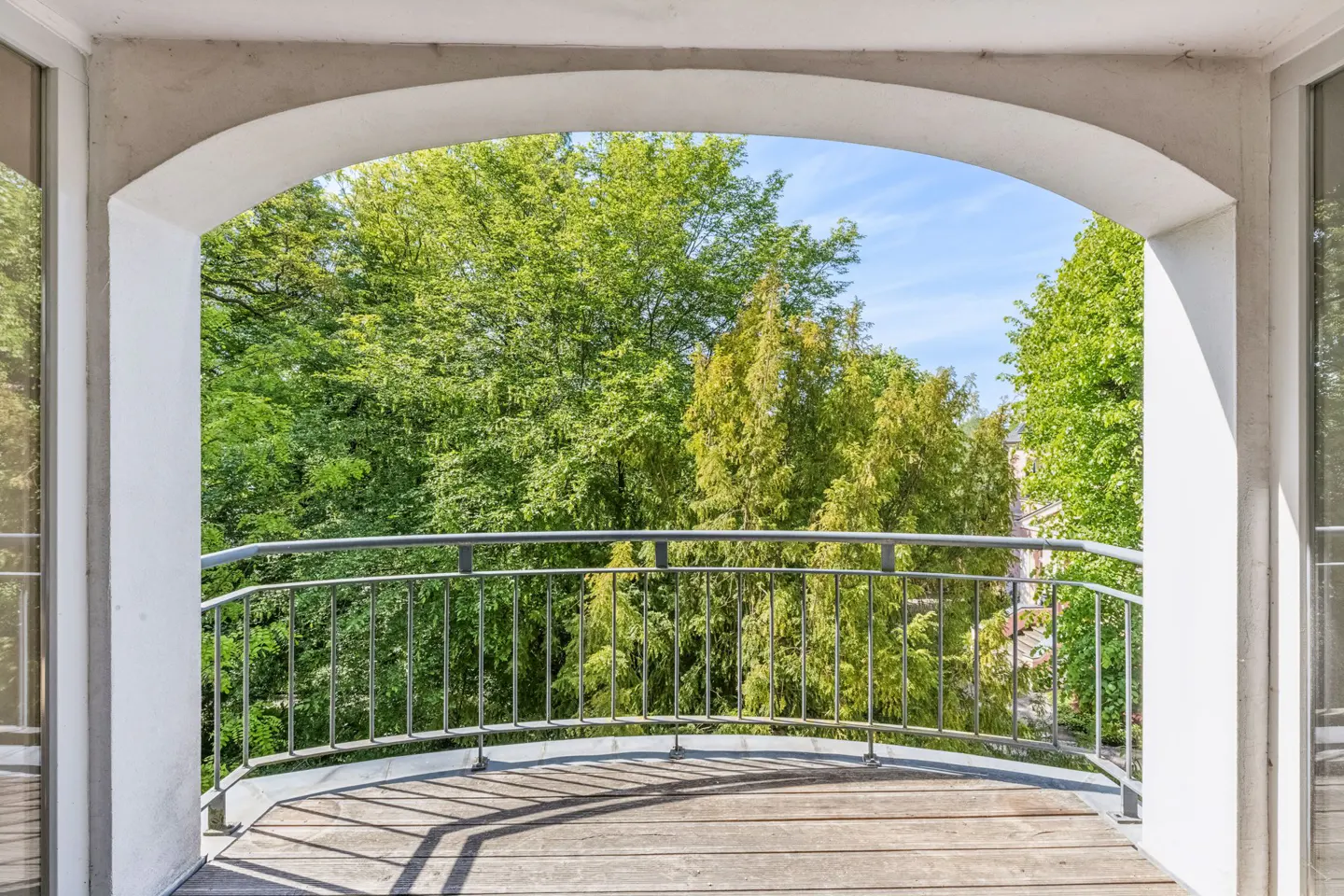 A curved balcony with a wooden floor and metal railing, framed by a white archway overlooking lush green trees.