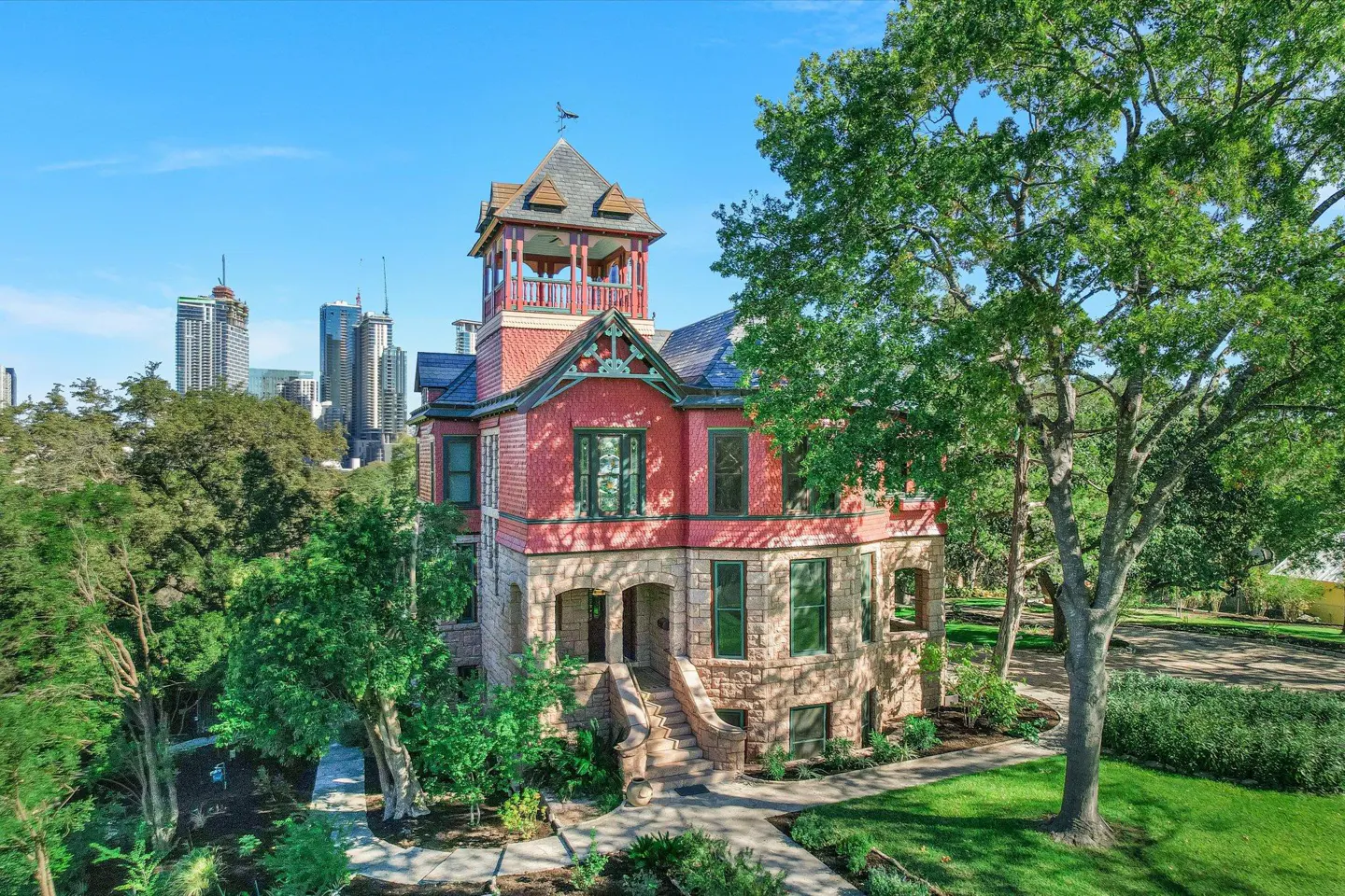 A three-story red and stone house with a tower, surrounded by green trees and grass, with city buildings in the background.