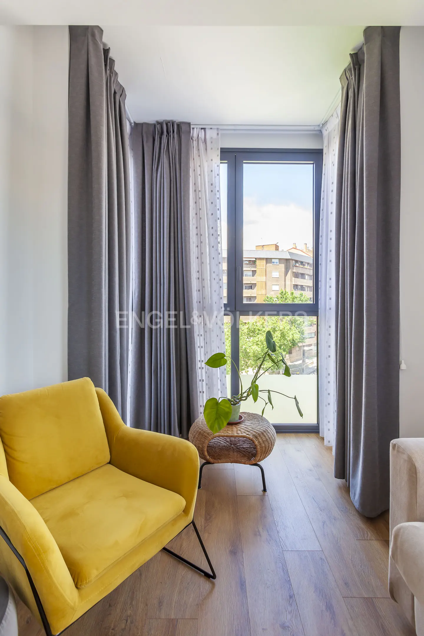 Bright living room with a yellow armchair, a round wicker coffee table with a plant, and grey curtains over a large window.