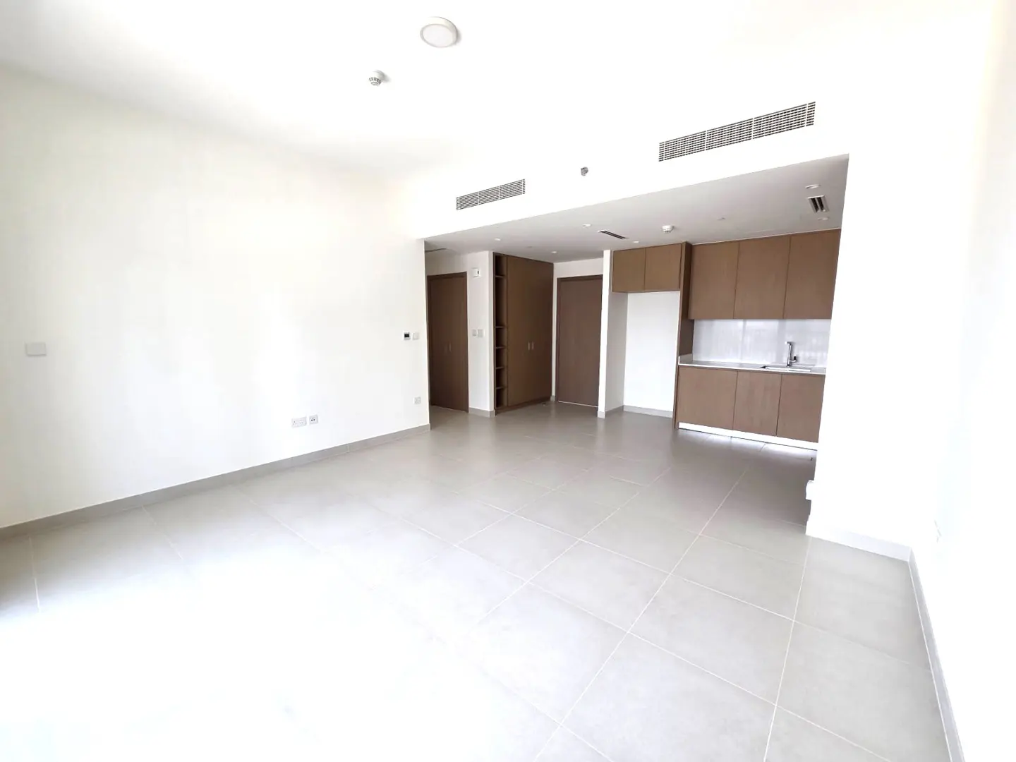 Bright, empty apartment with white walls and gray tile floor. Brown kitchen cabinets and doors are visible in the background.
