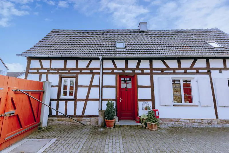 Two-story half-timbered house with a red door, white walls, and a gray tiled roof under a blue sky.